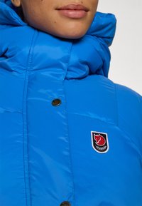 Blauwe pufferjack met een hoge kraag, voorzien van een opvallende logo patch en een gouden drukknoop. Glanzende, gewatteerde textuur.