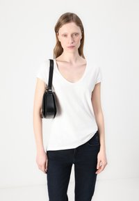 Camiseta blanca de cuello en V con mangas cortas, combinada con jeans azul oscuro. Un bolso negro cuelga del hombro. Textura de tela suave.