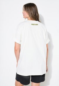 Hvid bomulds T-shirt med oprullede ærmer, prydet med grøn tekst "CHERRY DANCE" på den øverste del af ryggen. Afslappet pasform og glat tekstur.