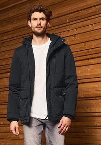 Černá prošívaná bunda s kapucí, s druky a zipem, předními kapsami a žebrovanými manžetami. Nositá přes bílé tričko.