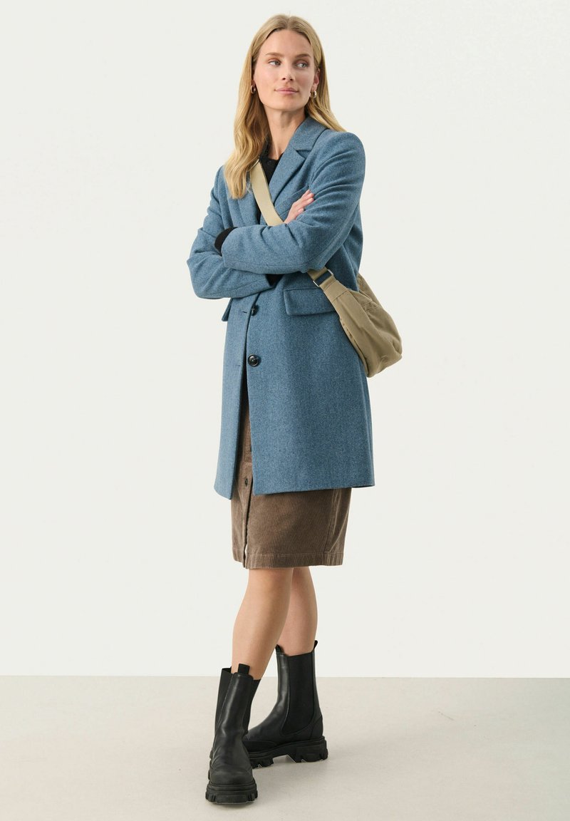 Niebieski płaszcz wełniano-syntetyczny o dopasowanym kroju, z zapięciem na jeden guzik i naszytymi kieszeniami; w połączeniu z brązową spódnicą z korduroy i czarnymi botkami za kostkę.
