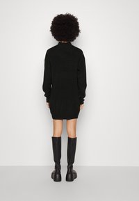 Robe noire en tricot à manches longues, texture côtelée et col montant. Portée avec des bottes noires montantes jusqu'aux genoux et un talon épais.