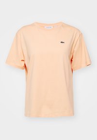 Lys ferskenfarvet bomulds t-shirt med løs pasform, rund halsudskæring og korte ærmer. Har et lille grønt broderet logo på øverste venstre bryst.