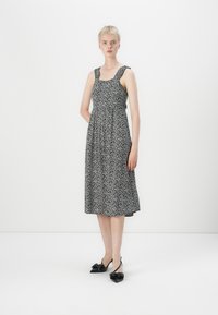 Μαύρο και άσπρο φόρεμα με λουλούδια, με τετράγωνη λαιμόκοψη και χοντρά λουριά, σε ρέουσα γραμμή Α, συνδυασμένο με μαύρες πέδιλες με τακούνι και φιόγκο.