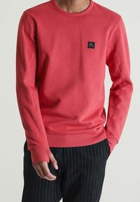 Sudadera coral con cuello redondo, puños y dobladillo acanalados, que presenta un pequeño parche de logo negro en el pecho. El material parece suave y texturizado.