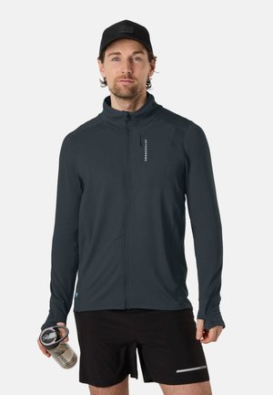 Mann mit schwarzer Kappe, dunkler Zip-Up-Jacke und schwarzen Shorts, der eine Wasserflasche hält und vor weißem Hintergrund steht.