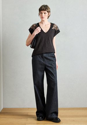 Sort kortærmet top med broderet blomsterdesign på skuldrene, kombineret med mørke denimbukser med vide ben. Modellen holder en lille sort taske.