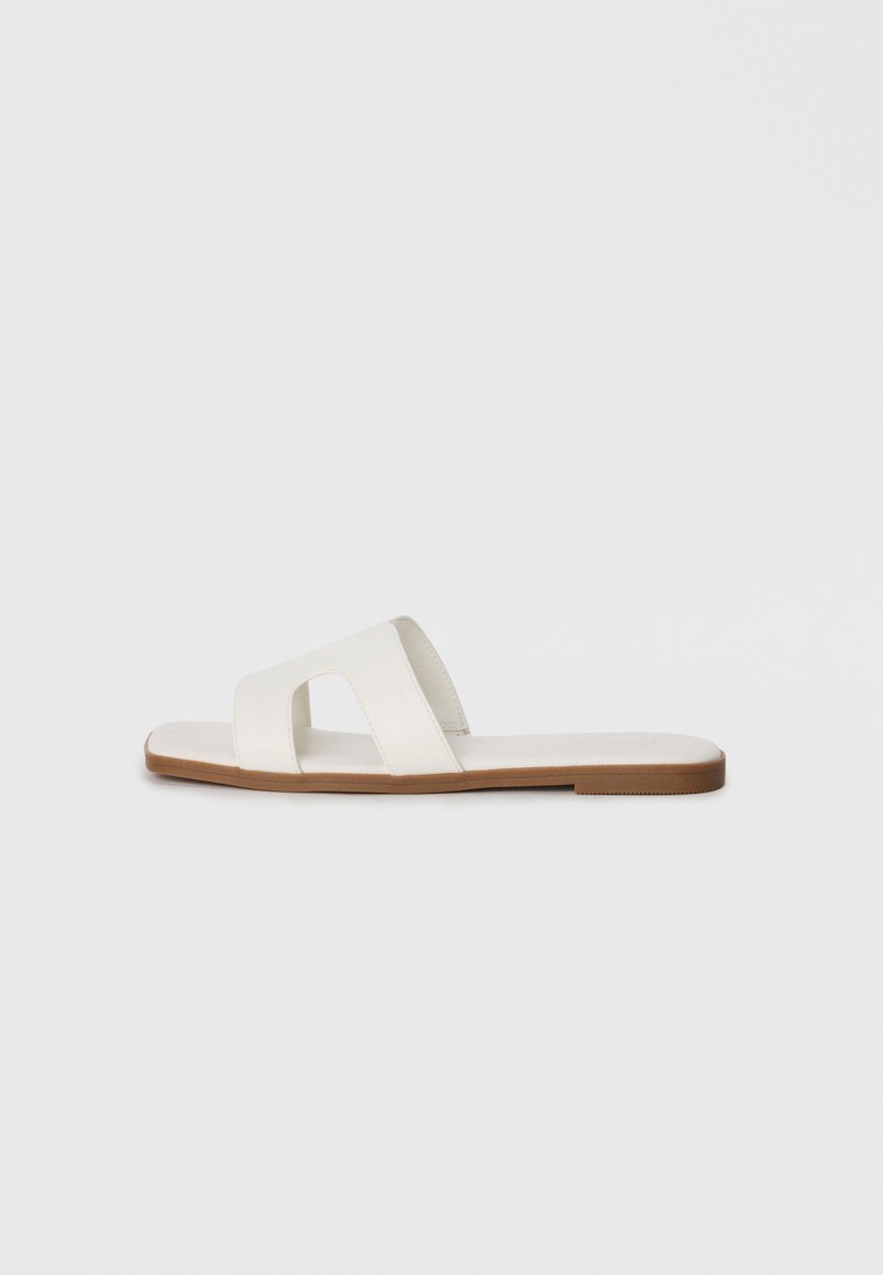 Sandale albe cu talpă plată, dotate cu două curele late cu decupaje, bază de culoare maro deschis, textură netedă și design minimalist.