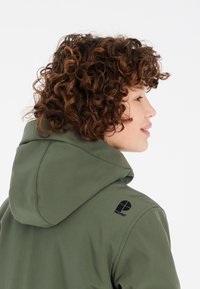 Veste à capuche verte en tissu doux avec une texture lisse. Elle présente un logo sur l'épaule et un design décontracté.