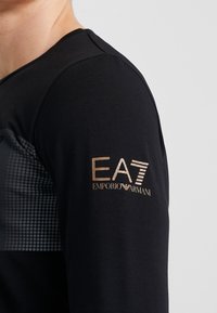 Svart långärmad skjorta med texturerat grått mönster på bröstet, med en gyllene "EA7 Emporio Armani"-logotyp på höger ärm.
