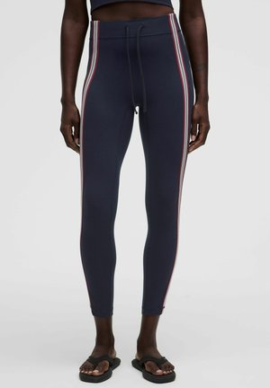 Slim fit marineblauwe legging met rode en witte strepen aan de zijkant, gedragen met zwarte slides bij een donkerhuidig persoon staand tegen een effen achtergrond.