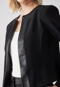 Black jacket with a tailored design, featuring a textured fabric and leather trim, round neckline, and fitted silhouette.