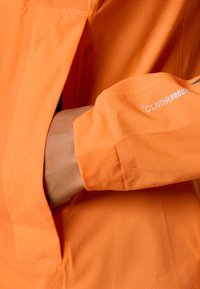 Oranger Jakker mit glatter Textur, nah an dem Bündchen, das das "CLIMAPROOF"-Markenzeichen zeigt. Sichtbare Nähte betonen das Design.