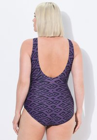 Vrouw met een blonde bobkapsel, gekleed in een paarse badpak met zwarte geometrische patronen, met haar rug naar een witte muur gekeerd.