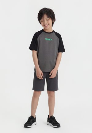 Jongen staand en glimlachend, gekleed in een grijs-zwart shirt met korte mouwen, donkergrijze korte broek en zwarte sportschoenen op een witte achtergrond.