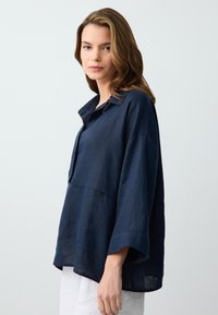 Camicia in lino blu navy con vestibilità ampia, grande colletto e maniche larghe, caratterizzata da una cucitura in vita e tessuto strutturato.