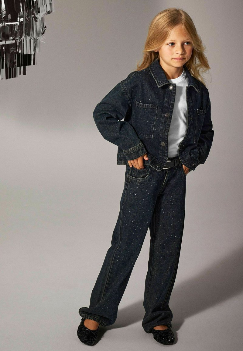 Mörk denimjacka och matchande jeans med metalliska dekorationer. Vit t-shirt under, tillsammans med svarta slip-on skor.