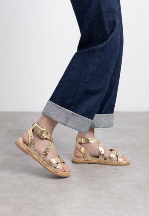 Sandalias de cuero dorado con detalles de tachuelas y una hebilla. La suela es de color marrón claro y las correas forman un diseño cruzado.