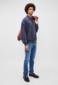 Pullover bleu închis cu fermoar și guler, asortat cu blugi bleu deschis și pantofi negri slip-on. Modelul ține o jachetă visinie.