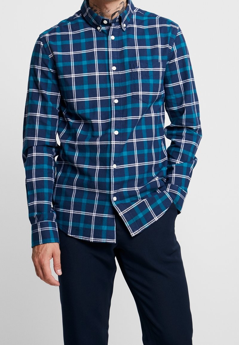 Chemise boutonnée arborant un motif à carreaux bleu et bleu sarcelle avec des accents blancs. Coupe ajustée, manches longues et détail d'une poche poitrine.