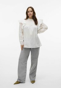 Blouse blanche avec des manches à volants, texture à pois et détails plissés, associée à un jean large gris et des chaussures vertes à bout pointu.