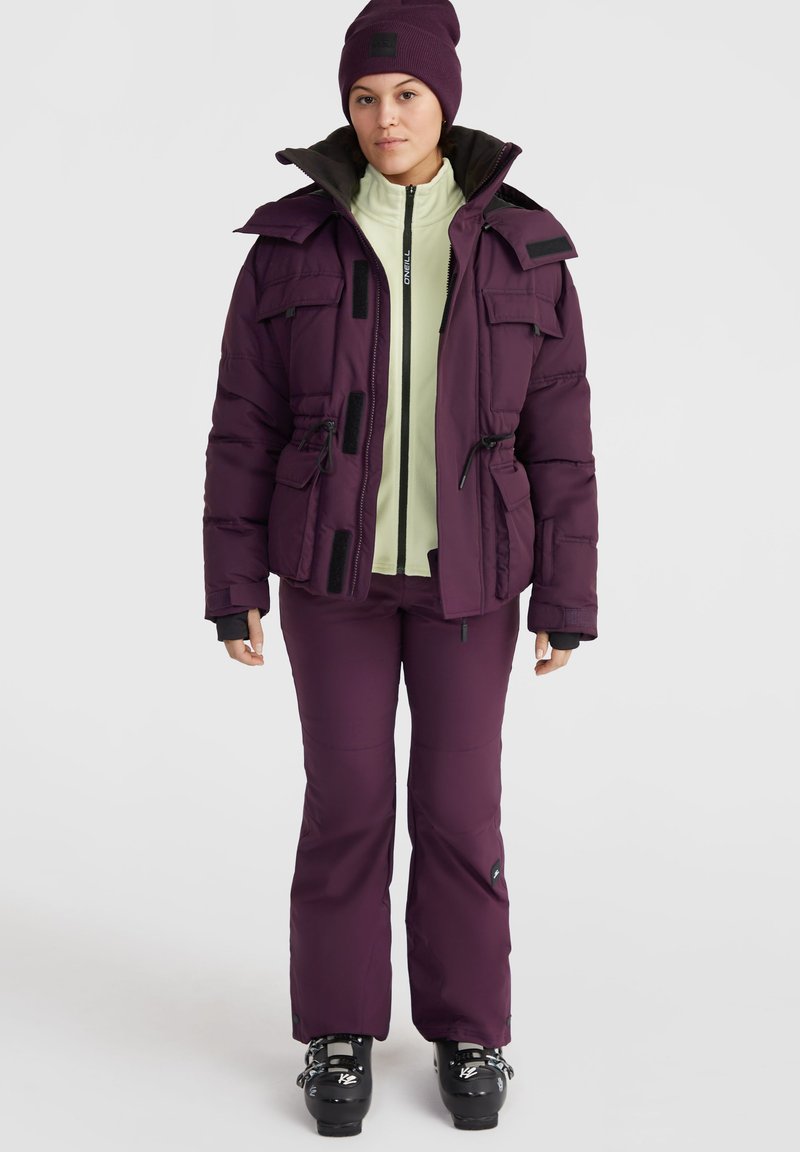 Daunenjacke in dunklem Lila, mit hohem Kragen, Taschen und Kordelzug. Kombiniert mit einem hellgrünen Fleecepullover und passenden lila Hosen.