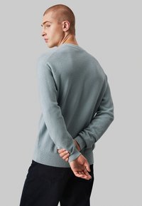 Lichtblauwe gebreide trui met een textuurpatroon, ronde halslijn, geribbelde manchetten en zoom. Model staat iets van je af, met de handen achter de rug.