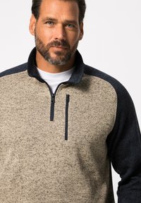 Pull en beige et marine avec un col zippé et un tissu texturé ; présente des épaules contrastantes et une petite poche zippée sur la poitrine.