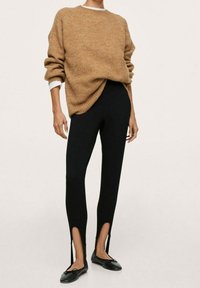 Brązowy, oversize sweter wykonany z miękkiego materiału, w połączeniu z czarnymi dopasowanymi spodniami z rozcięciami przy kostkach oraz czarnymi płaskimi butami.