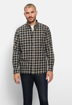 Mann mit kurzen Haaren, der ein braunes, blaues und weißes kariertes Hemd über einem weißen Unterhemd und dunklen Jeans trägt, vor hellem Hintergrund.