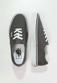 Grå canvas sneakers med vita skosnören, rund tå och strukturerad yta. Svart kant längs sulan och Vans logotyp på inlägget.
