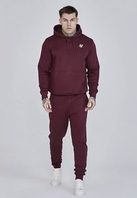 Vinröd hoodie med framficka och dragsko, kombinerad med matchande joggingbyxor. Har logga på bröstet och sidan. Slät tygstruktur.
