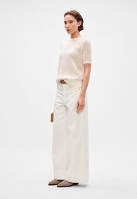 Svetlý béžový kašmírový sveter s krátkymi rukávmi, skombinovaný s bielymi nohavicami so širokými nohavicami a hladkými semišovými lodičkami v pieskovej farbe. Minimalistický dizajn s voľným strihom.