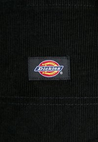 Černá žebrovaná látka se čtvercovou nášivkou loga Dickies ve středu, která obsahuje červenou, žlutou a bílou barvu.