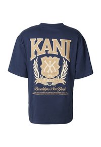 Tamsiai mėlynas medvilninis marškinėliai su dideliu šviesiai rudos spalvos grafinu, kuriame yra "KANI", šarvo emblema ir tekstas apie Brukliną, Niujorką. Trumpi rankovės.