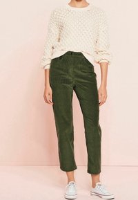 Leichtes, crèmefarbenes Strickpullover mit strukturiertem Muster, kombiniert mit olivgrünen Cordhosen im geraden Schnitt, getragen von einem Modell mit weißen Sneakers.