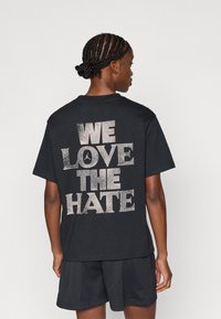 Μαύρη βαμβακερή μπλούζα T-shirt με γυαλιστερό λευκό κείμενο στην πλάτη που γράφει "ΜΑΣ ΑΡΕΣΕΙ ΤΟ ΜΙΣΟΣ," με στρογγυλή λαιμόκοψη και κοντά μανίκια.