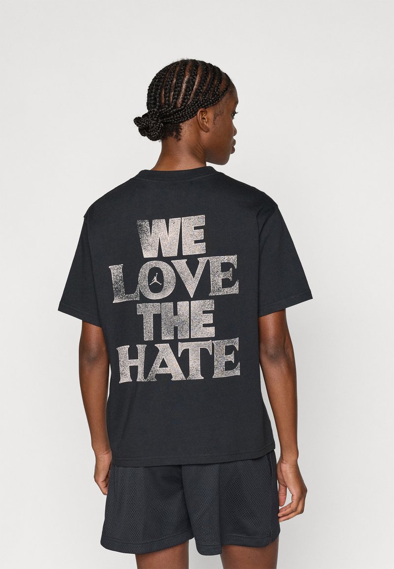 Μαύρη βαμβακερή μπλούζα T-shirt με γυαλιστερό λευκό κείμενο στην πλάτη που γράφει "ΜΑΣ ΑΡΕΣΕΙ ΤΟ ΜΙΣΟΣ," με στρογγυλή λαιμόκοψη και κοντά μανίκια.