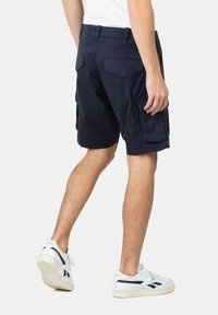 Pantalones cortos cargo azul marino con dos bolsillos laterales, dos bolsillos traseros y una cinturilla. Llevados con zapatillas blancas con detalles en azul.