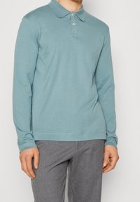 Pánská polokošile s dlouhým rukávem v světle modré barvě z bavlny, má klasický límec a tříknoflíkovou légu, doplněna šedými kalhotami. Hladký materiál.