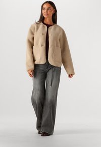 Beige, gestructureerde jas met een ronde halslijn, twee voorzakken en houten knopen; gecombineerd met grijze wijde jeans.