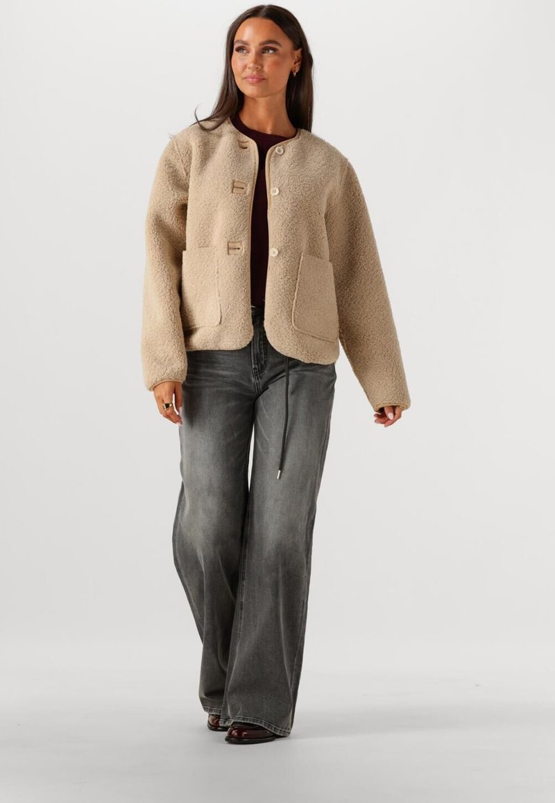 Beige, gestructureerde jas met een ronde halslijn, twee voorzakken en houten knopen; gecombineerd met grijze wijde jeans.