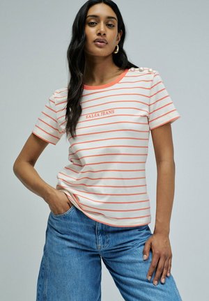 Joven mujer con cabello largo y oscuro viste una camisa blanca con rayas coral con el texto "SALSA JEANS" y vaqueros azules, mano en el bolsillo, fondo neutro.