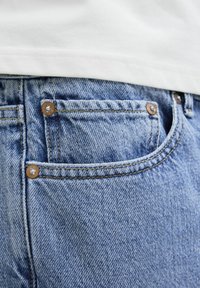 Jeans de mezclilla en azul claro con una vista en primer plano del bolsillo trasero. Presentan remaches de cobre y costuras en contraste. Textura suave.