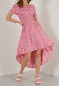 Vaaleanpunainen olkaimeton mekko, jossa on korkea-matala helma, valkoisella kukkakuviolla, lyhyet hihat ja istuva bodice. Päällä valkoiset sandaalit.