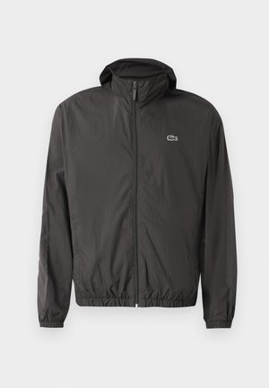 Giacca leggera nera con cappuccio, polsini elasticizzati e chiusura con zip. Presenta un piccolo logo ricamato sul lato sinistro del petto. Texture liscia.