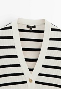 Cardigan en maille blanche avec des rayures horizontales noires, col en V et un seul bouton rond doré à l'avant.