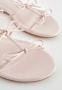 Vaaleanpunaiset litteät sandaalit, jotka on valmistettu saumattomasta nahasta. Niissä on koristeelliset solmittavat hihnat ja minimalistinen muotoilu, jossa on pyöreä kärki.