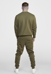 Olivgrön tvådelad träningsoverall med sweatshirt och avsmalnande byxor. Tyget verkar mjukt med ribbade muddar och avslappnad passform.