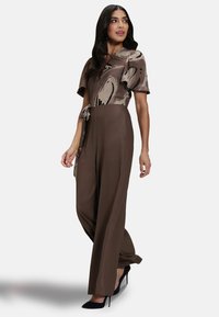 Brown jumpsuit with a patterned short-sleeve top, cinched waist, and wide-leg pants. Features a tie accent at the waist and black high heels.